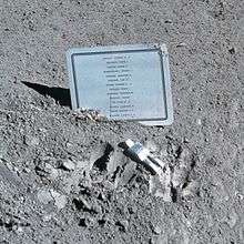 A silvery human figure, apparently face down, in front of a metallic plaque with a list of names on a dusty gray surface