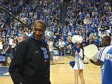 A man, wearing a black jacket and black T-shirt, is walking onto the basketball court.