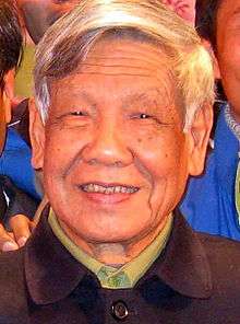 An old graying man wearing a brown buttoned-up jacket, and a pale green shirt