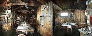 Dimly lit interior of a timber structure, with (left) a long table on which are papers and other unidentifiable objects. There are also bunks, sheles and packing cases. To the right, under a small window, is a smaller table with a chair.